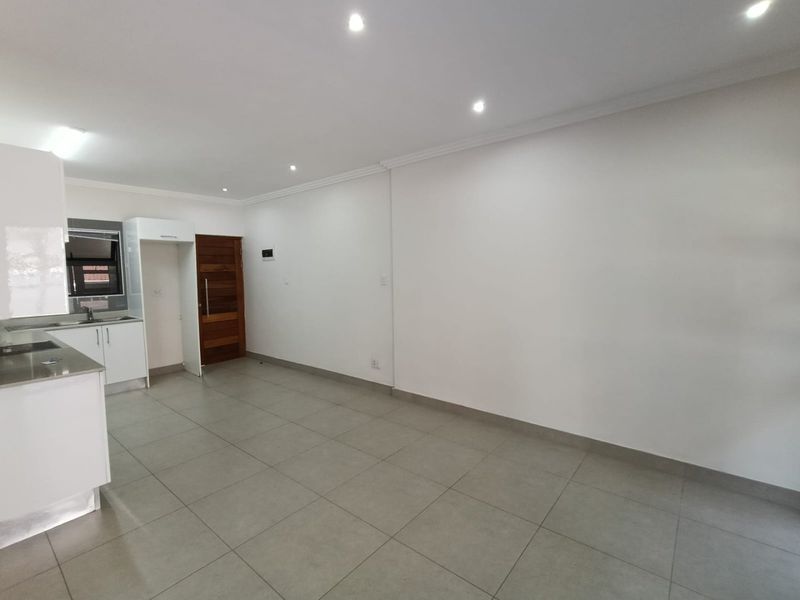 A bright and spacious open-plan living area featuring modern tiled flooring, recessed downlighting, and a compact kitchen with white cabinetry.