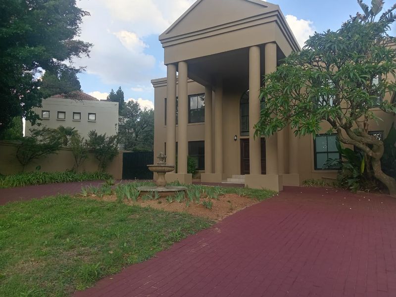 A grand double-story residence featuring a prominent portico with columns, a paved driveway, and a central stone fountain in the front garden.