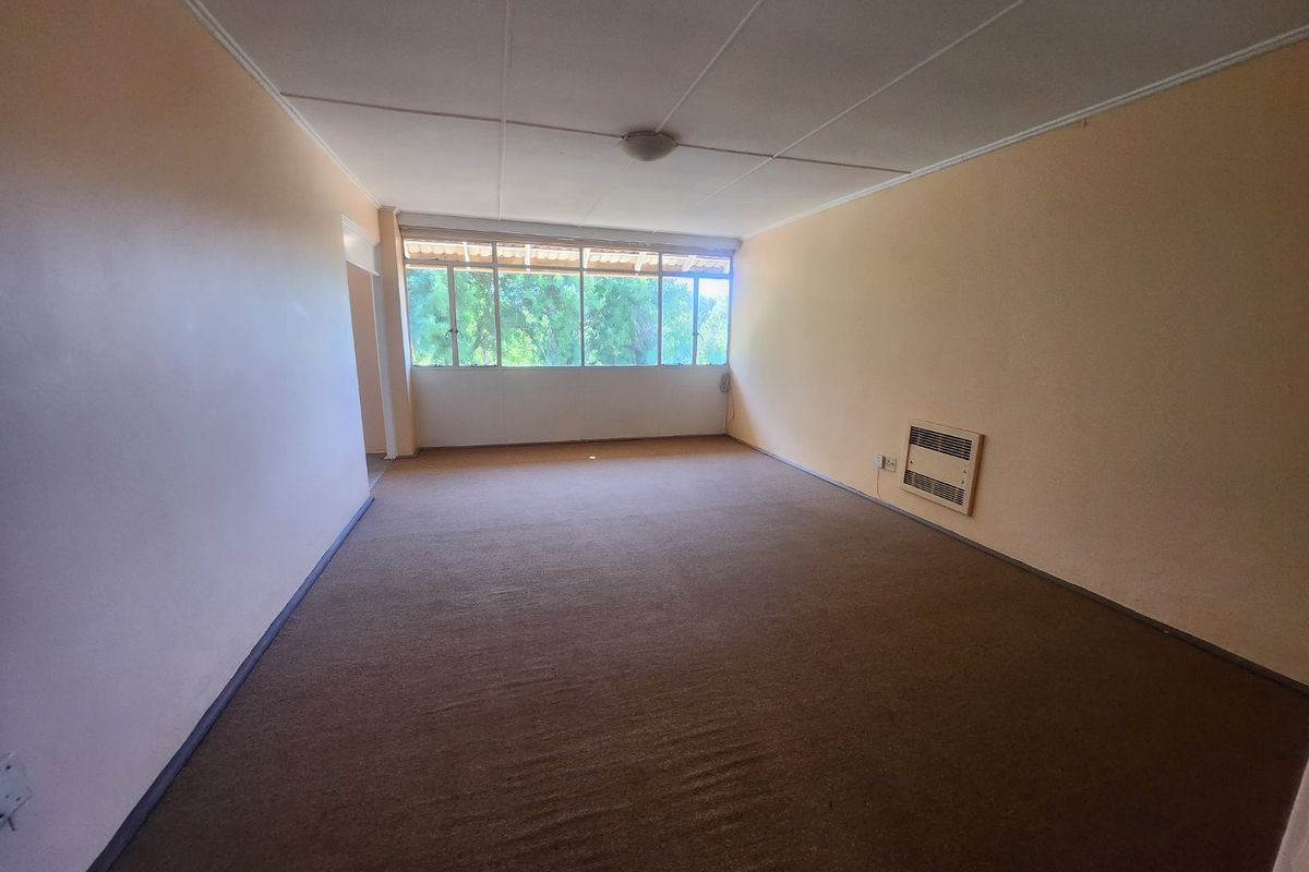 A spacious, empty living room featuring wall-to-wall carpeting, a wall-mounted air conditioning unit, and large windows providing natural light.