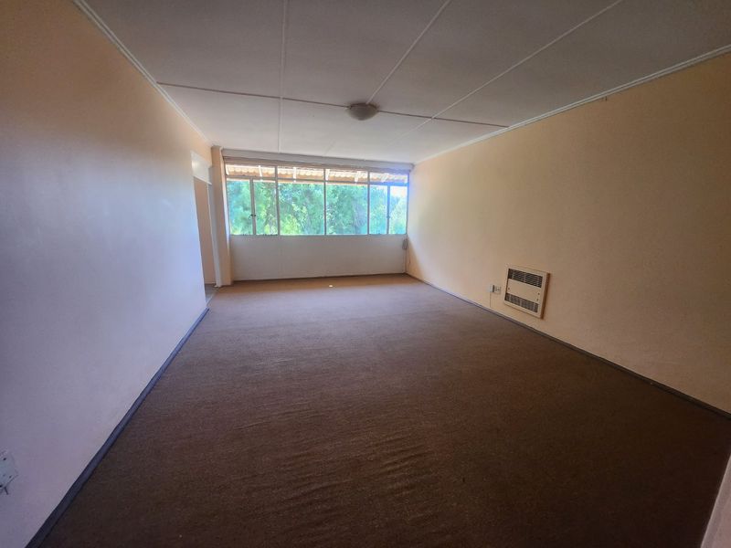 A spacious, empty living room featuring wall-to-wall carpeting, a wall-mounted air conditioning unit, and large windows providing natural light.