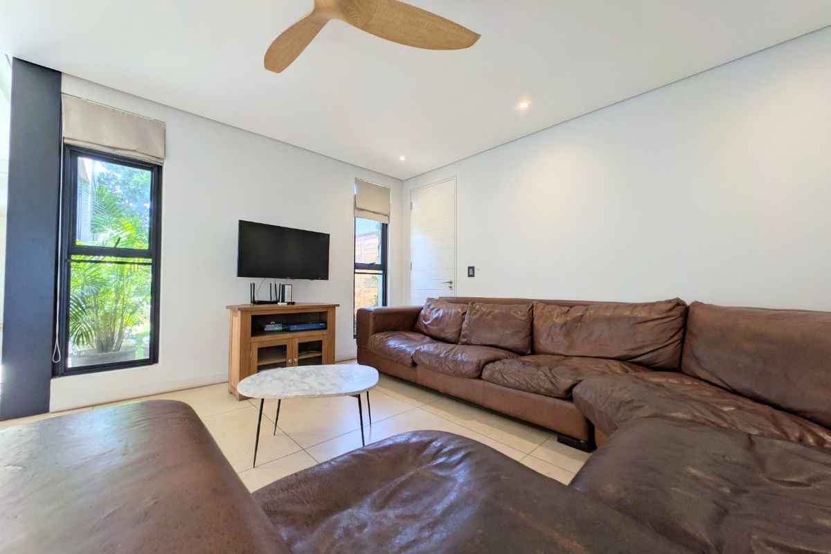 A bright and modern living room featuring tiled flooring, recessed downlights, and a contemporary ceiling fan, with large windows providing natural light.