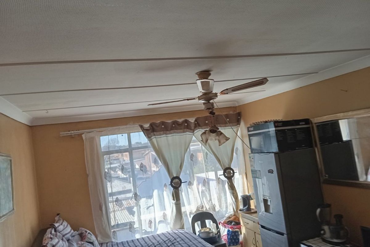 A bedroom featuring a ceiling fan and large window, showing signs of wear on the ceiling and walls.