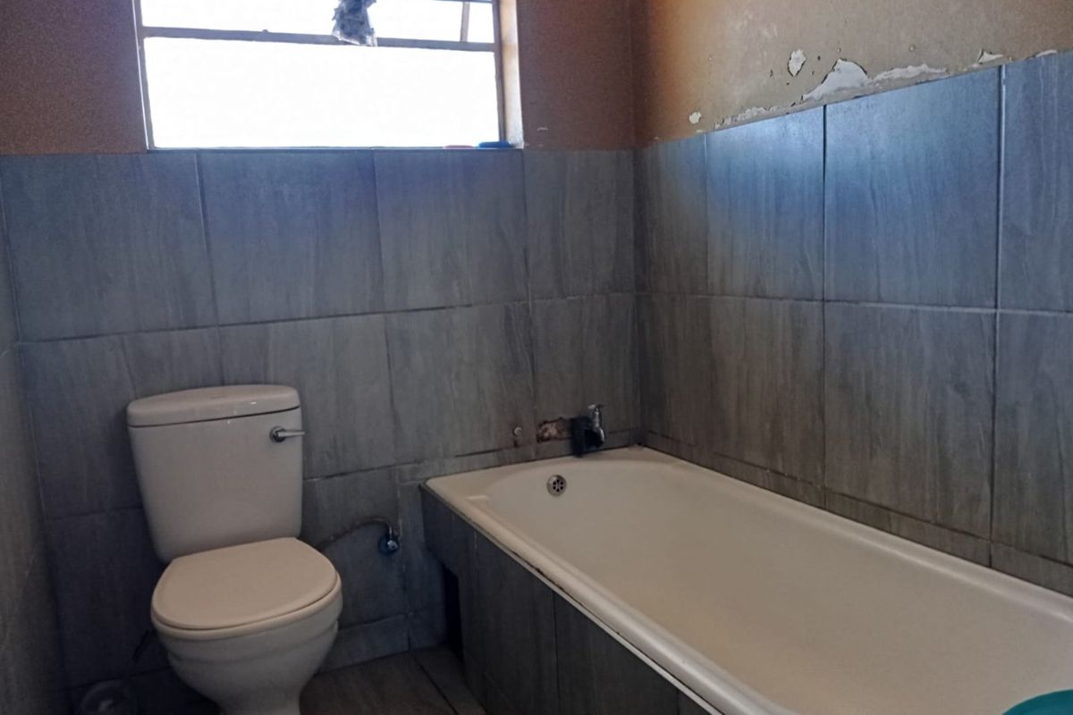 A bathroom featuring a bathtub and toilet with grey wall tiling. The space requires maintenance and cosmetic repairs to the wall finishes.
