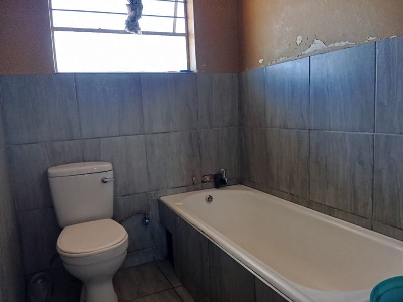 A bathroom featuring a bathtub and toilet with grey wall tiling. The space requires maintenance and cosmetic repairs to the wall finishes.