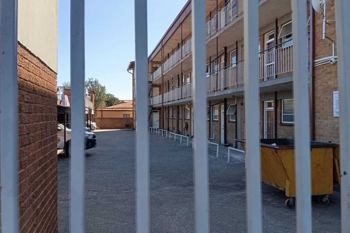 View of the apartment complex rear exterior, featuring a paved parking area, brick facade, and metal security fencing.