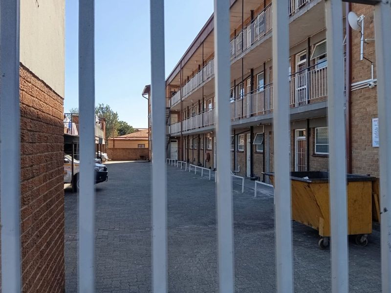 View of the apartment complex rear exterior, featuring a paved parking area, brick facade, and metal security fencing.