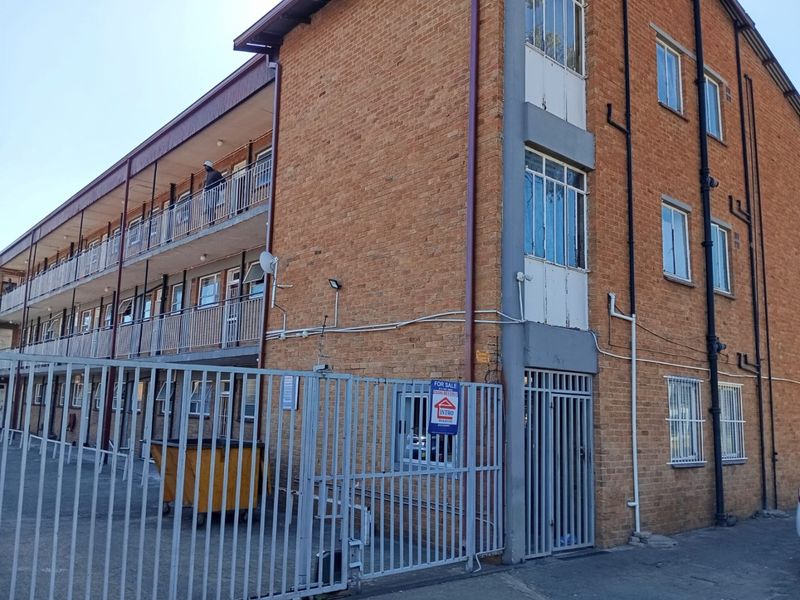 Exterior view of a multi-story brick apartment building featuring metal security fencing, balcony access, and visible window frames.