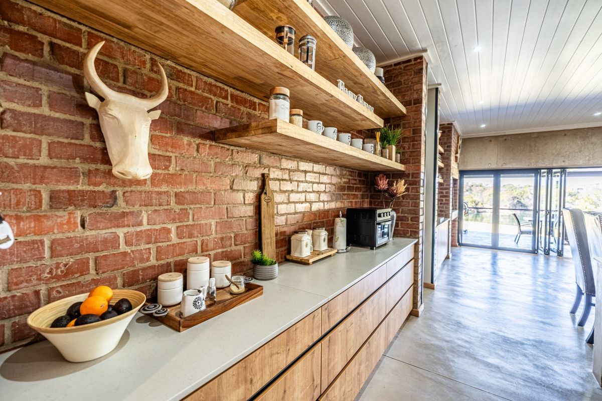 Modern kitchen featuring exposed brick walls, timber floating shelves, stone benchtops, and sleek timber cabinetry, leading to an open-plan living area with bi-fold doors.
