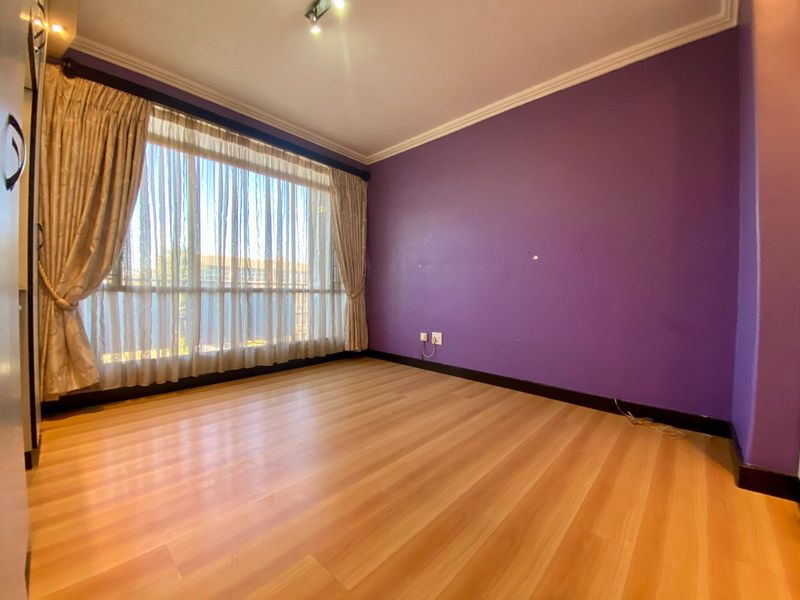 An empty bedroom featuring laminate flooring, built-in cupboards, and large windows with curtains. The room requires minor wall repairs and a fresh coat of paint.