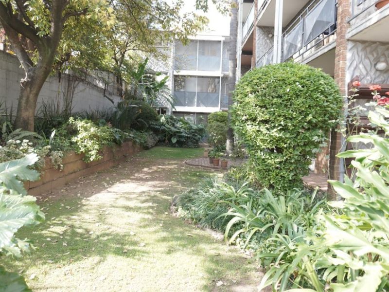 A landscaped garden area featuring a retaining wall, mature trees, and established shrubs, adjacent to the apartment building's balcony structure.