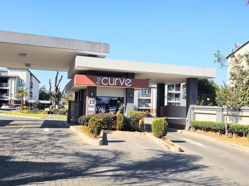 The secure entrance gatehouse of The Curve apartment complex, featuring a paved driveway, boom gate access, and perimeter security fencing.