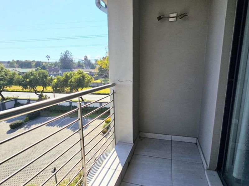 A compact balcony featuring tiled flooring, a stainless steel balustrade, and a wall-mounted light fixture, offering an elevated view of the parking area.