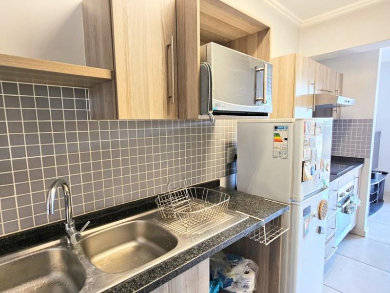 A functional kitchen featuring light wood-tone cabinetry, dark laminate countertops, a double stainless steel sink, and a tiled backsplash, leading into an open living area.
