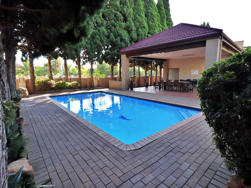 A well-maintained outdoor pool area featuring a paved deck, mature trees, and a covered patio structure for shaded relaxation.