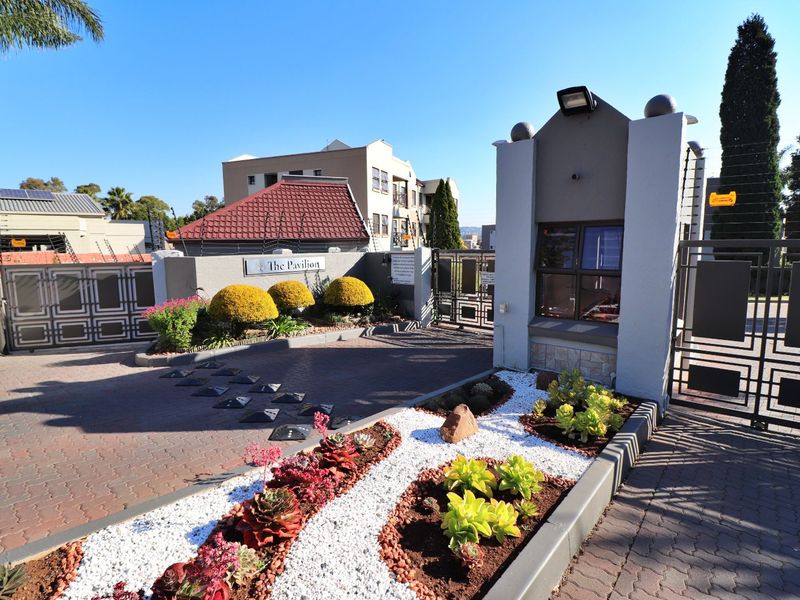 The secure entrance to The Pavilion apartment complex, featuring a guard house, automated security gates, electric fencing, and well-maintained landscaped garden beds.