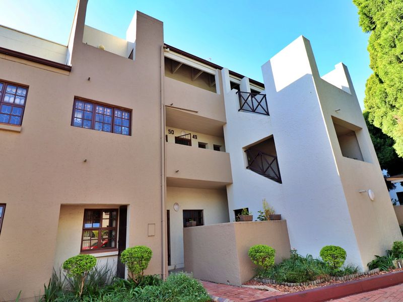 Front elevation of a multi-story apartment complex featuring balconies, security window bars, and established garden beds with brick paved pathways.