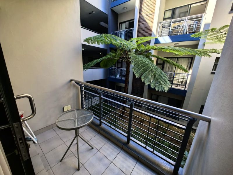 A private apartment balcony featuring tiled flooring and a metal balustrade, overlooking a central courtyard with established greenery.