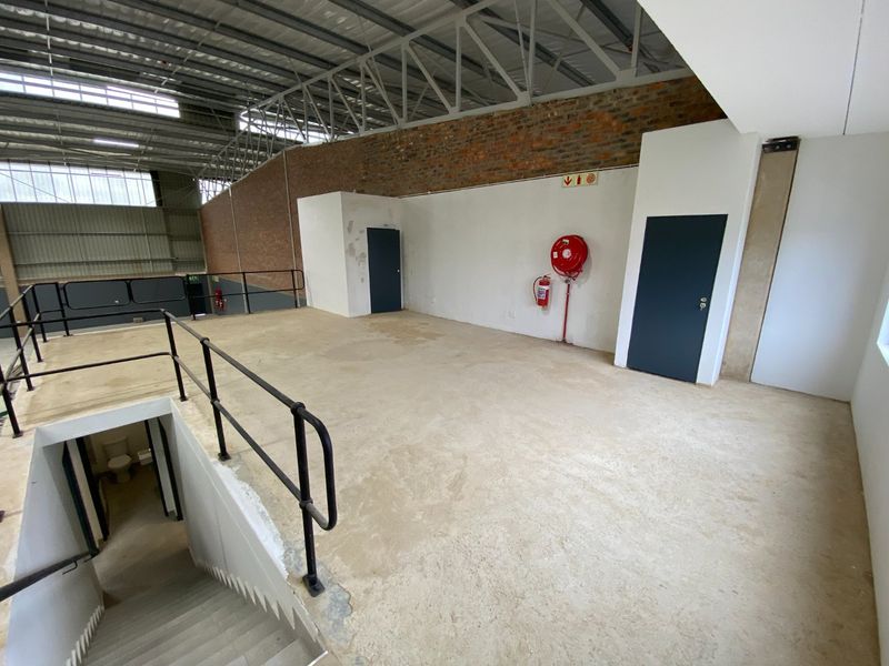 An open-plan industrial mezzanine level featuring exposed steel trusses, brickwork, and essential fire safety equipment, accessible via a concrete staircase.