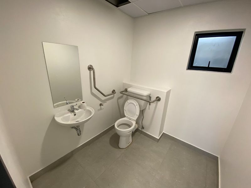 A clean, functional accessible bathroom featuring a wall-mounted basin, toilet, and safety grab rails with neutral tiled flooring.