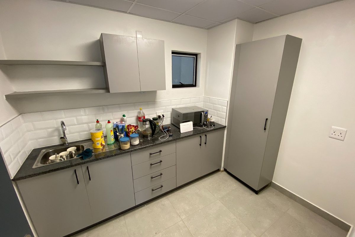 A clean, modern kitchenette featuring grey cabinetry, a dark countertop, subway tile backsplash, and a tall pantry unit in an industrial office setting.