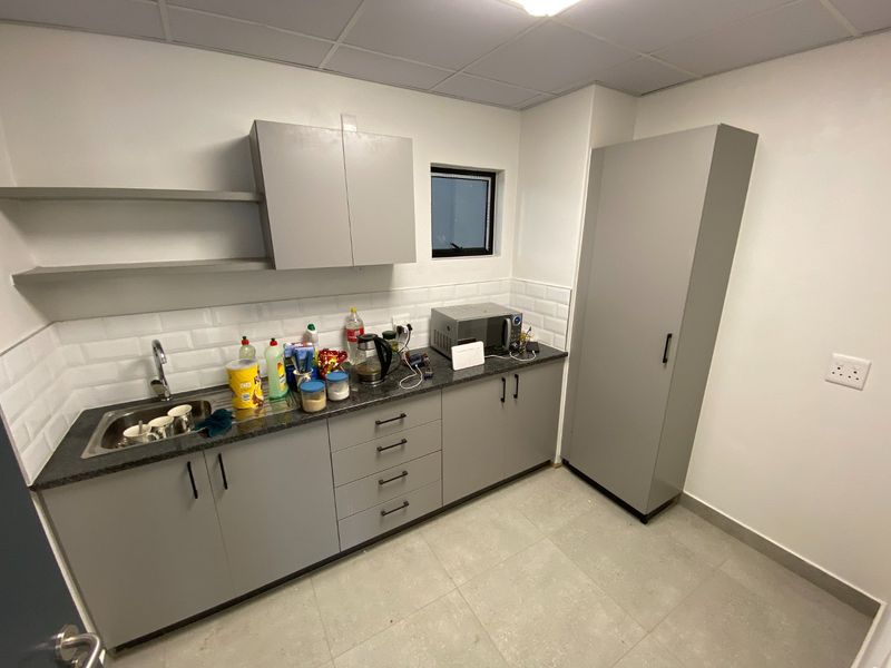 A clean, modern kitchenette featuring grey cabinetry, a dark countertop, subway tile backsplash, and a tall pantry unit in an industrial office setting.