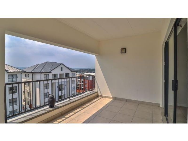 A spacious covered balcony featuring tiled flooring, a metal balustrade, and sliding glass door access, offering an open view of the surrounding apartment complex.