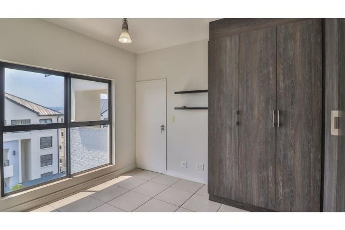 A bright, empty bedroom featuring modern tiled flooring, built-in dark wood-look cupboards, and large aluminium-framed windows offering natural light.