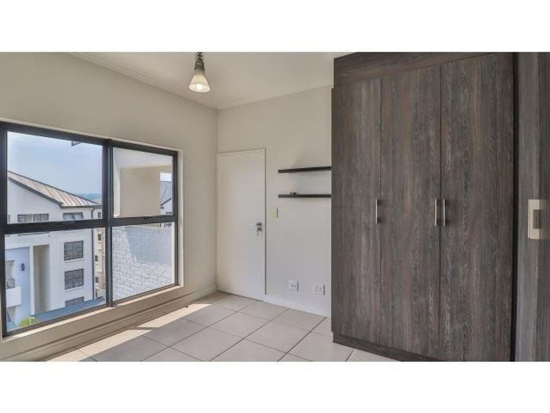 A bright, empty bedroom featuring modern tiled flooring, built-in dark wood-look cupboards, and large aluminium-framed windows offering natural light.