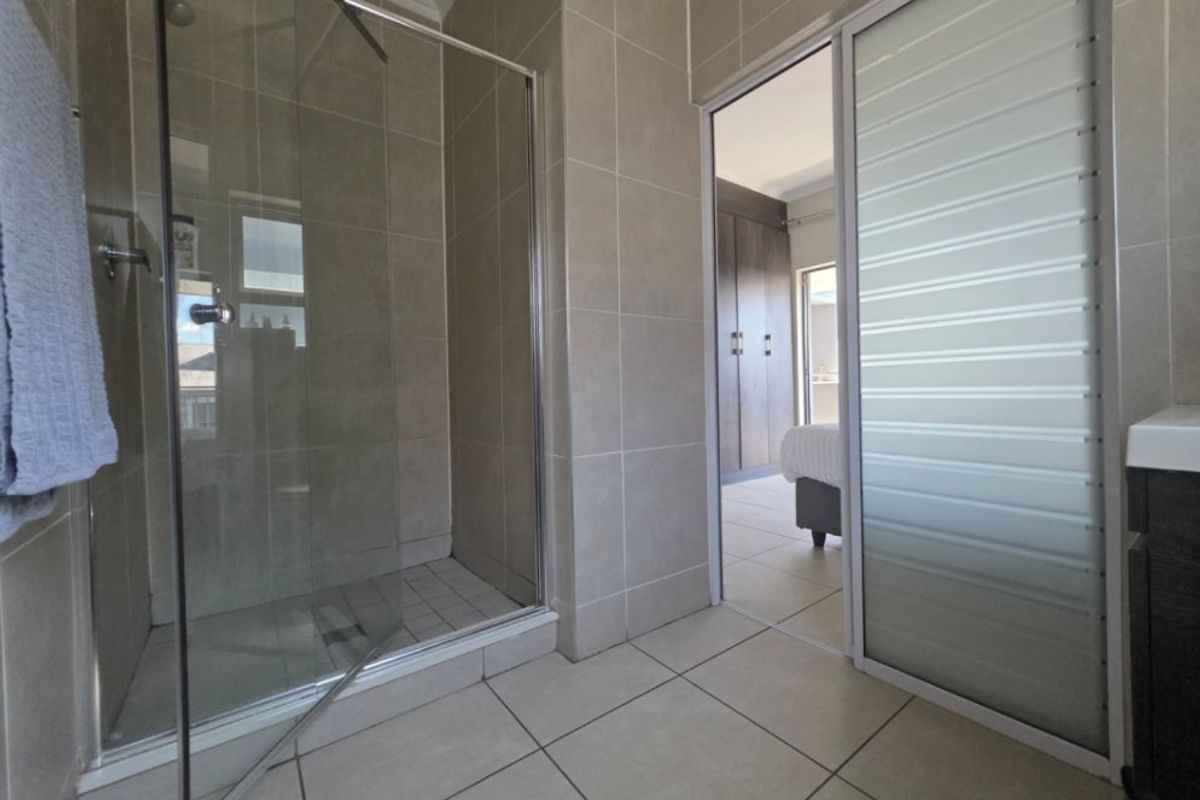 A modern bathroom featuring a glass-enclosed shower, neutral wall and floor tiling, and a sliding door providing access to the adjacent bedroom.