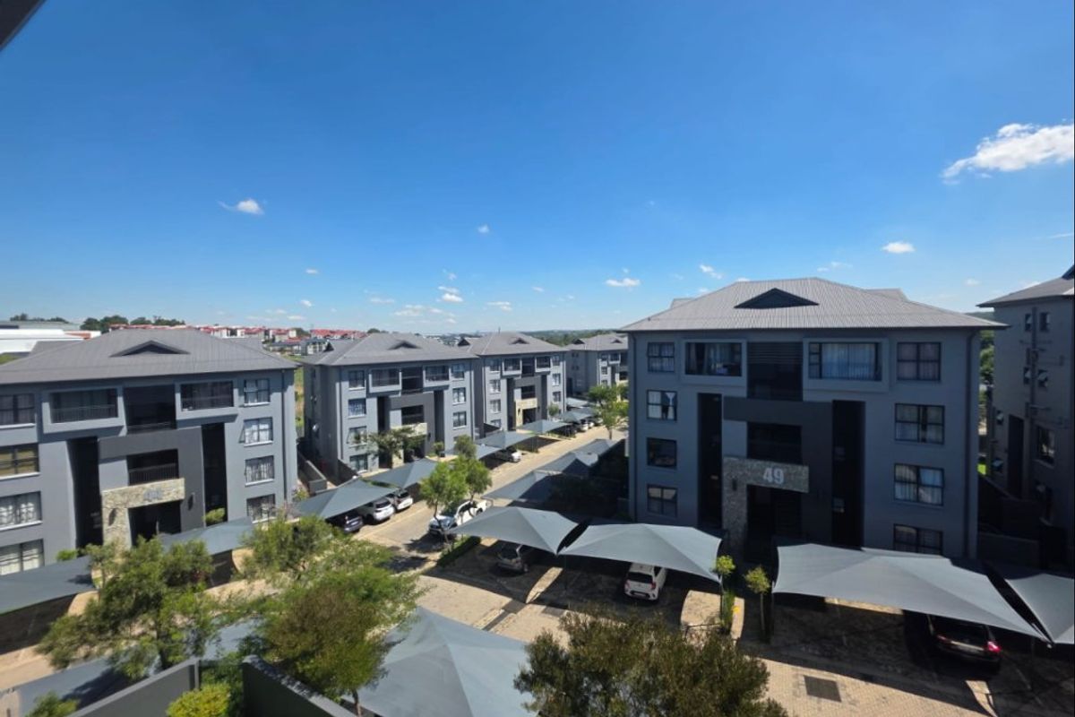 An elevated aerial view of a modern apartment complex featuring multiple residential blocks, paved driveways, and covered shade cloth parking bays.