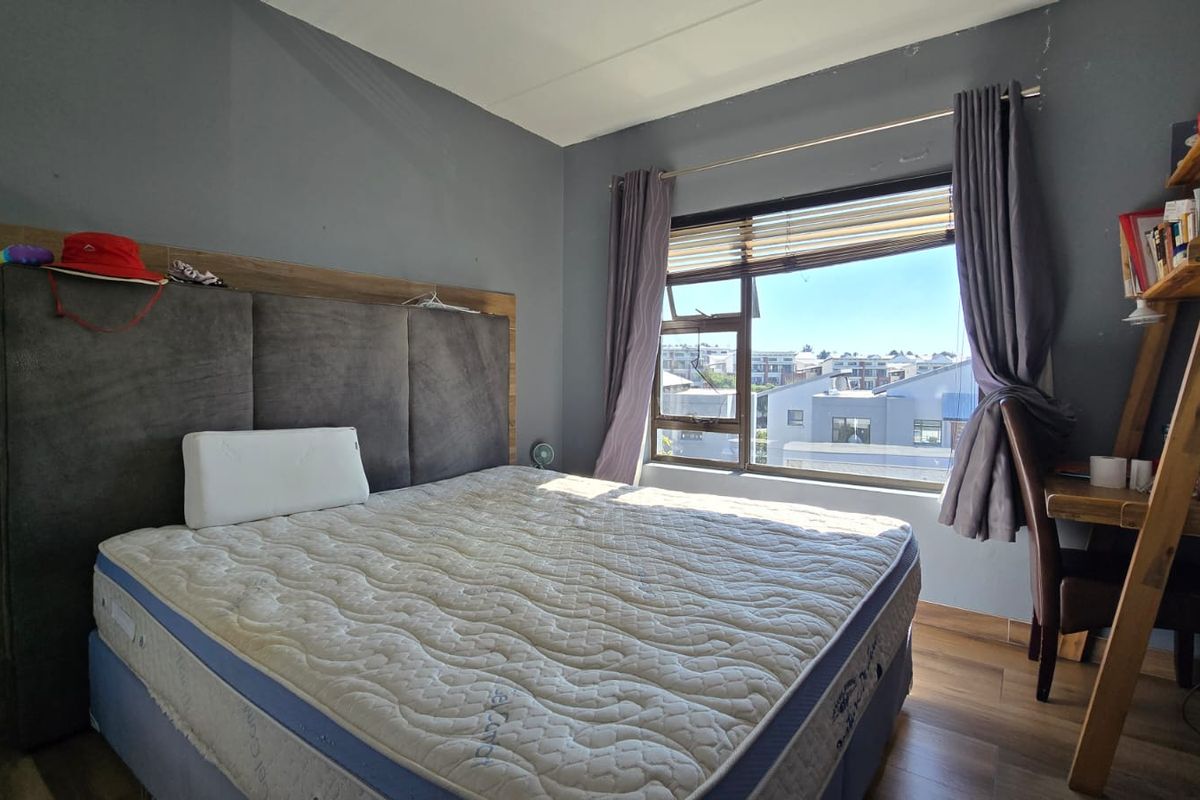 A bedroom featuring laminate flooring, an upholstered headboard, and a window with blinds offering an elevated view of the surrounding neighborhood.