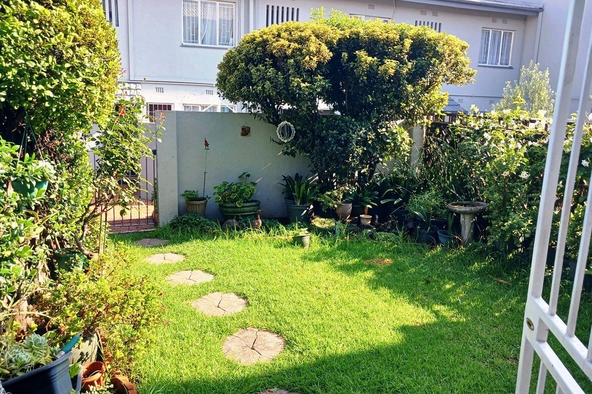 A private rear garden area featuring a lawn, established greenery, stepping stones, and a secure boundary wall with a metal gate.