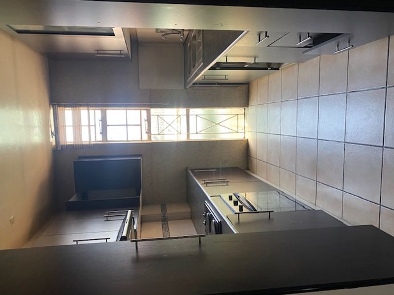 A galley-style kitchen featuring dark built-in cabinetry, laminate countertops, integrated oven and hob, and tiled flooring with a window providing natural light.