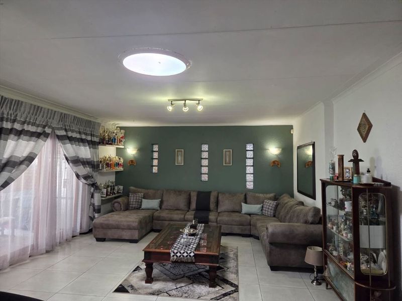 A spacious living room featuring tiled flooring, a green accent wall with decorative glass block inserts, and recessed ceiling lighting.