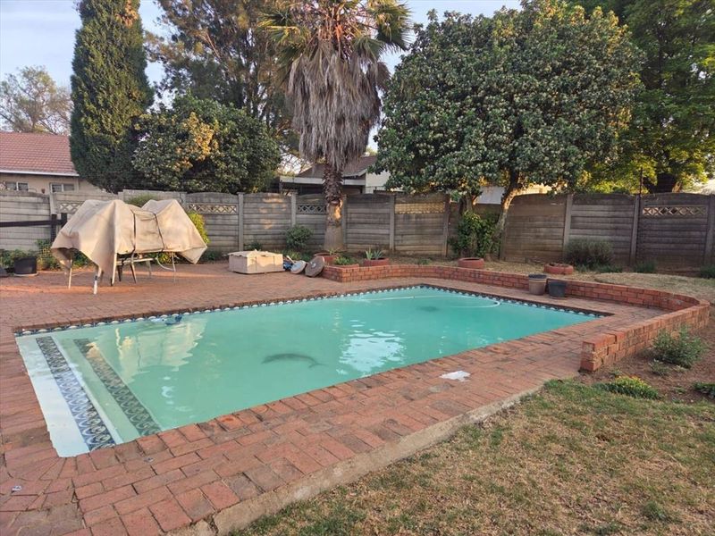 A backyard pool area featuring a rectangular swimming pool with brick paving surround and a concrete boundary wall.
