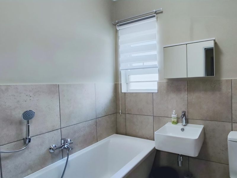 A modern bathroom featuring a bathtub with handheld shower, wall-mounted basin, toilet, and a mirrored medicine cabinet with neutral wall tiling.