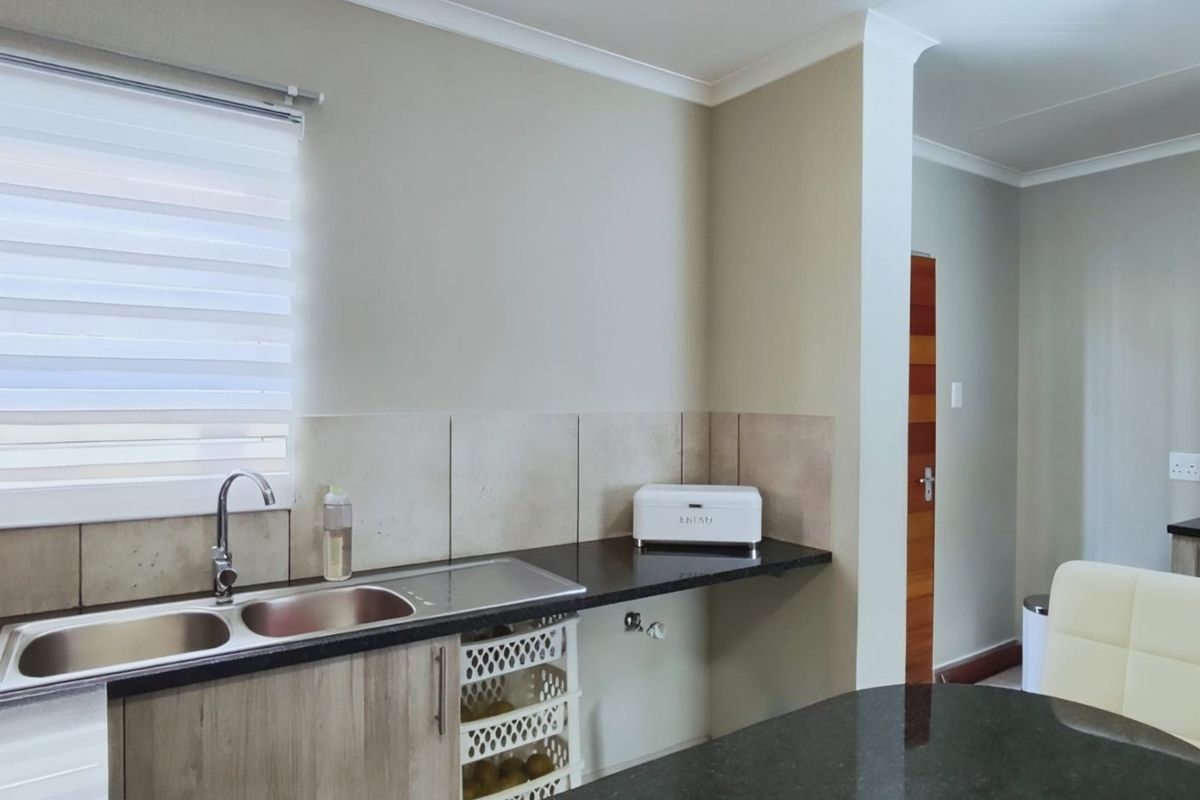 A modern kitchen area featuring granite countertops, a double sink, tiled backsplash, and neutral cabinetry with integrated storage.
