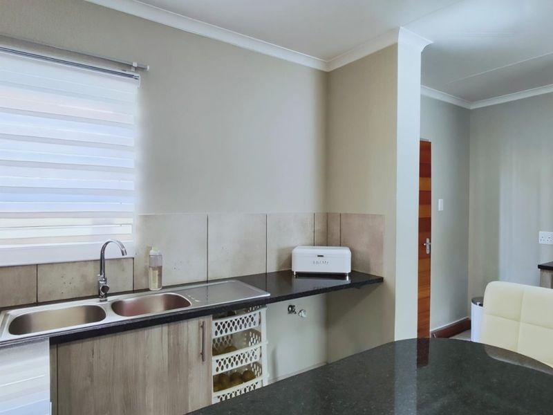 A modern kitchen area featuring granite countertops, a double sink, tiled backsplash, and neutral cabinetry with integrated storage.