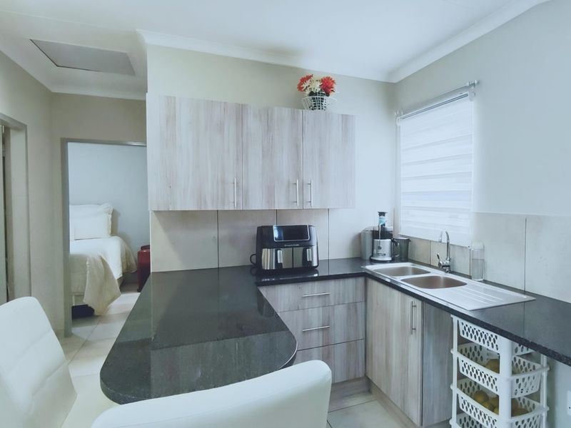 A functional kitchen featuring light wood-look cabinetry, dark granite countertops, a double sink, and a tiled backsplash with a view into an adjacent bedroom.