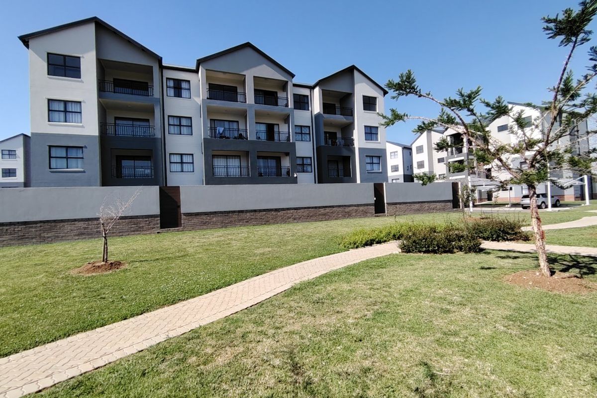 Exterior view of a modern apartment complex featuring multi-story residential blocks with private balconies, a paved walkway, and well-maintained communal garden areas.