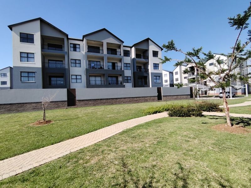 Exterior view of a modern apartment complex featuring multi-story residential blocks with private balconies, a paved walkway, and well-maintained communal garden areas.