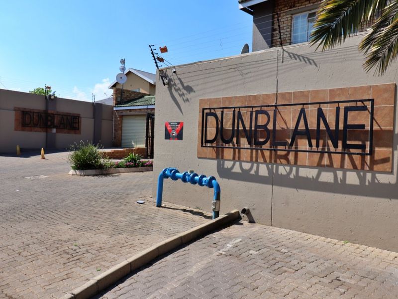 Entrance view of the Dunblane complex featuring a paved driveway, perimeter wall with signage, and security infrastructure.