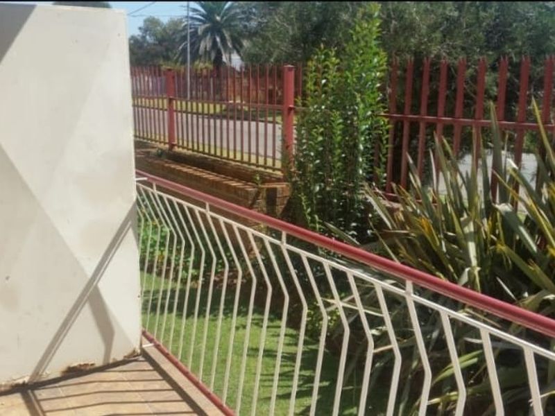 A small balcony area featuring tiled flooring and a metal balustrade, overlooking a garden space with security fencing.