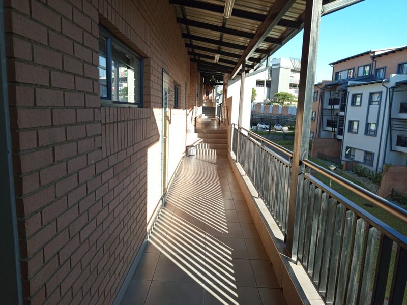 A covered outdoor walkway or balcony featuring tiled flooring, brick walling, and a metal balustrade overlooking the apartment complex grounds.