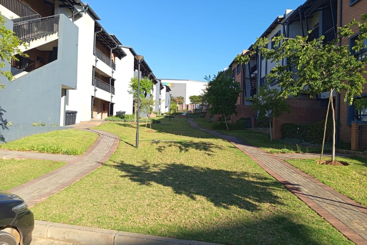 A well-maintained communal garden area between apartment blocks featuring paved walkways, manicured lawns, and established trees within a residential complex.