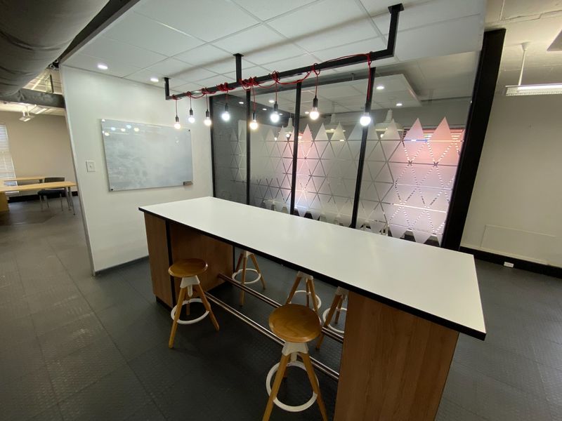 Modern office breakout area featuring a built-in high-top table, glass partition wall with frosted detailing, and contemporary pendant lighting.
