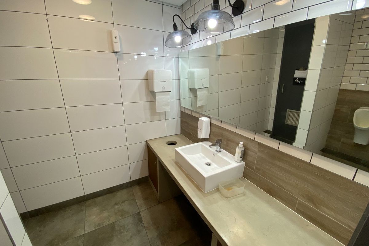 A clean, modern commercial bathroom featuring a wall-mounted basin, large vanity mirror, tiled surfaces, and contemporary wall-mounted lighting.