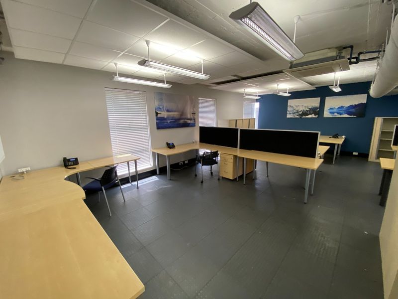 Open-plan commercial office space featuring suspended ceilings, industrial-style exposed ducting, durable rubber flooring, and large windows with venetian blinds.