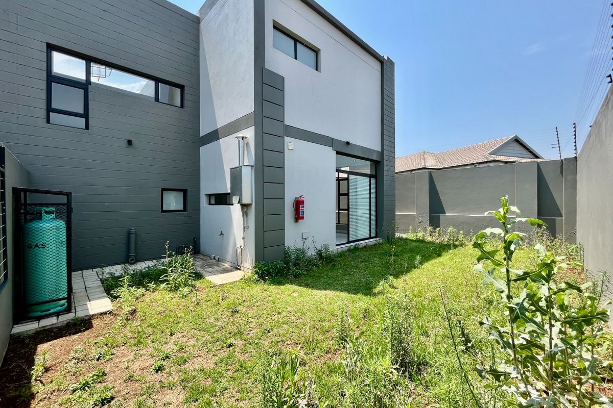 Rear exterior view of a modern two-story house featuring a paved walkway, gas cylinder storage, and a private garden area with boundary walling.