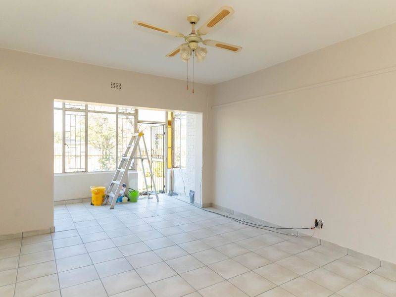 An empty, open-plan living area featuring tiled flooring, a ceiling fan, and large windows with metal security bars.